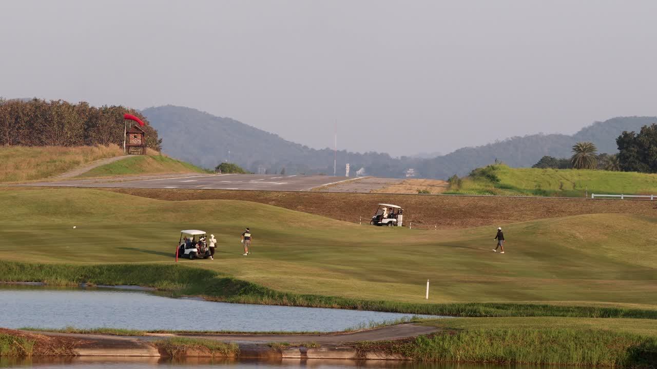 Golfers and carts on lush fairway by lake, mountains behind, warm sunset lighting