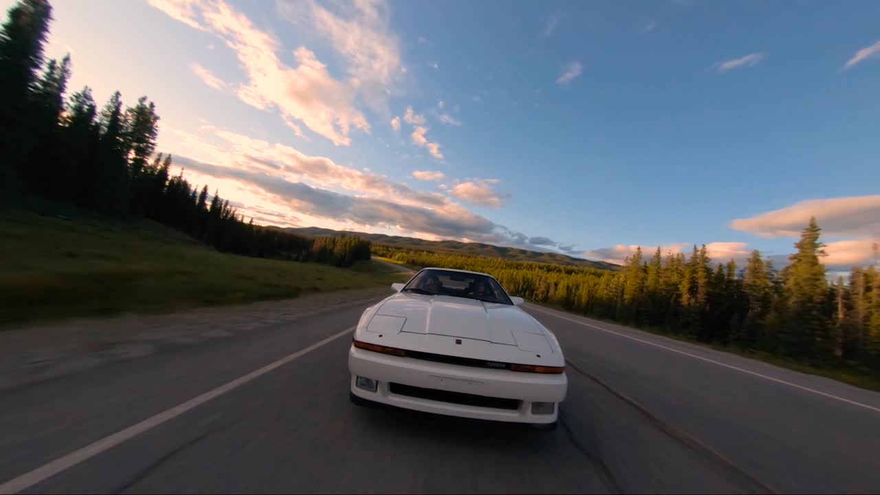 A white sports car seen pulling up fast to a drone. The timing is perfect and the sky is filled with color from the setting sun.