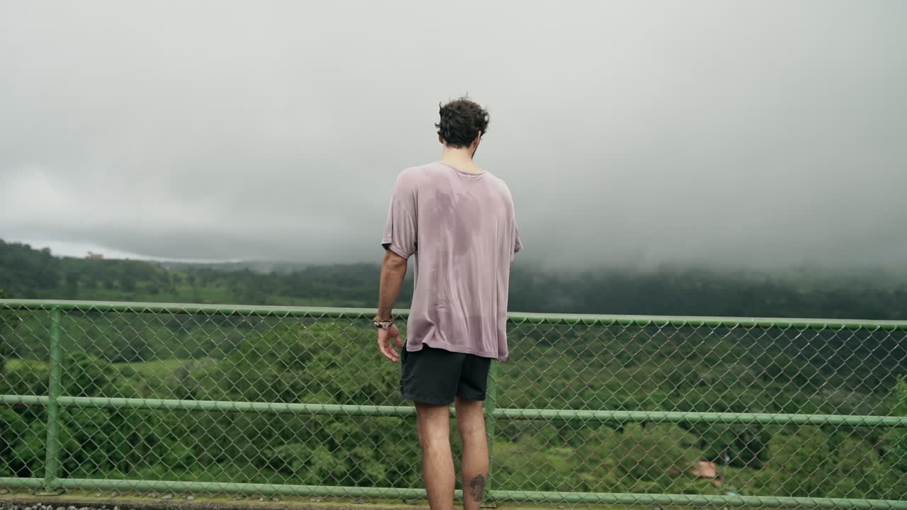 Man stares over railing at overlook as shirt blows in wind, slow motion