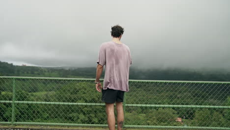 El-Hombre-Mira-Por-Encima-De-La-Barandilla-Al-Mirador-Mientras-La-Camisa-Sopla-En-El-Viento,-Cámara-Lenta