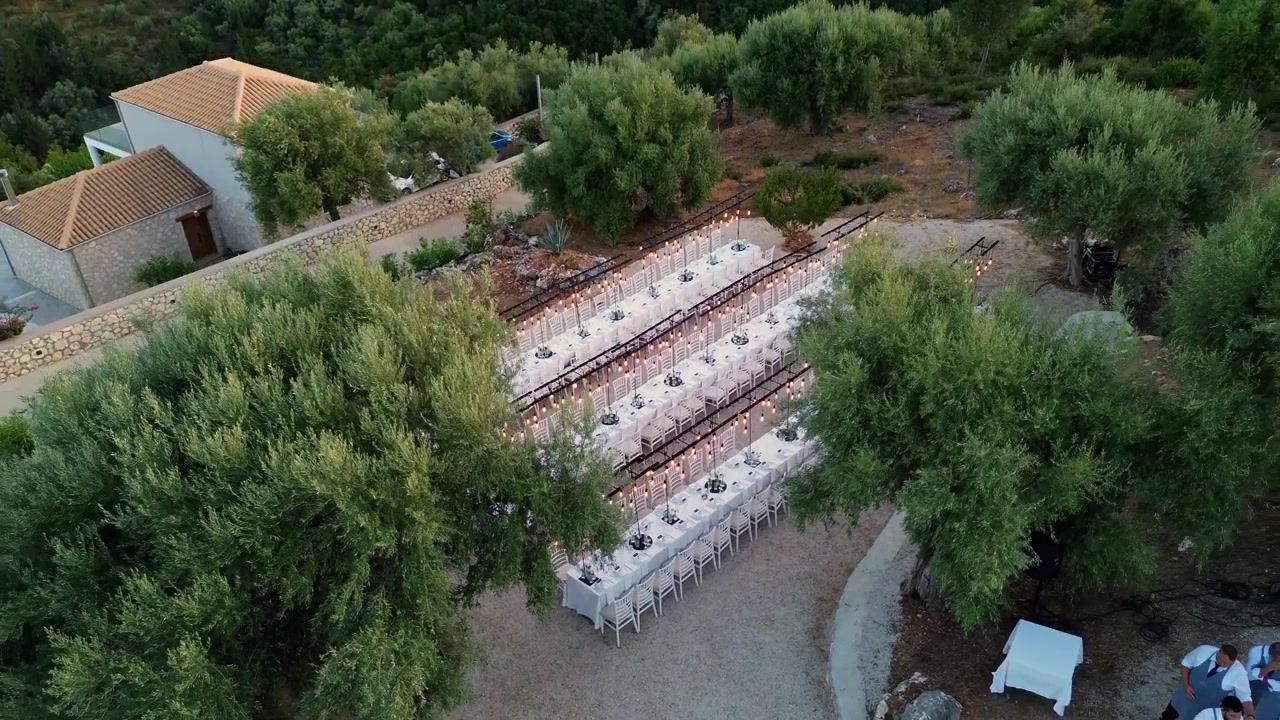 Cinematic pull-away drone shot flying backward to reveal long decorated wedding banquet tables surrounded by beautiful green olive trees.