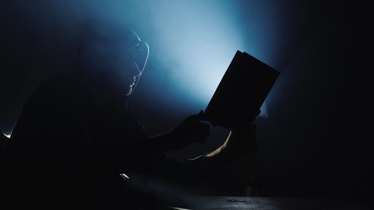 силуэт человека в капюшоне с книгой в руках от книги исходит свет