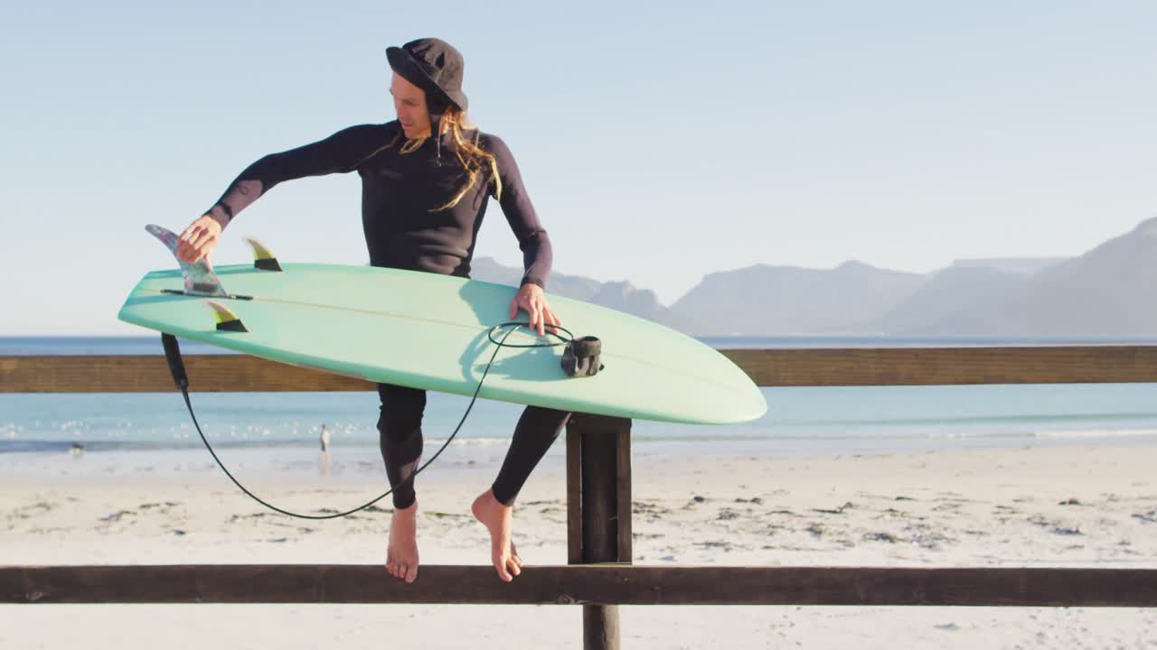video de un hombre caucásico con dreadlocks en traje de neopreno comprobando la tabla de surf sentado en el paseo marítimo
