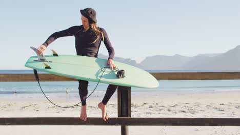 video of caucasian man with dreadlocks in wetsuit checking surfboard sitting on beach promenade