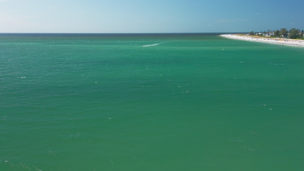 Turquoise waters stretch along a sandy shoreline with boats cruising offshore, as a coastal town lines the horizon under a bright blue sky