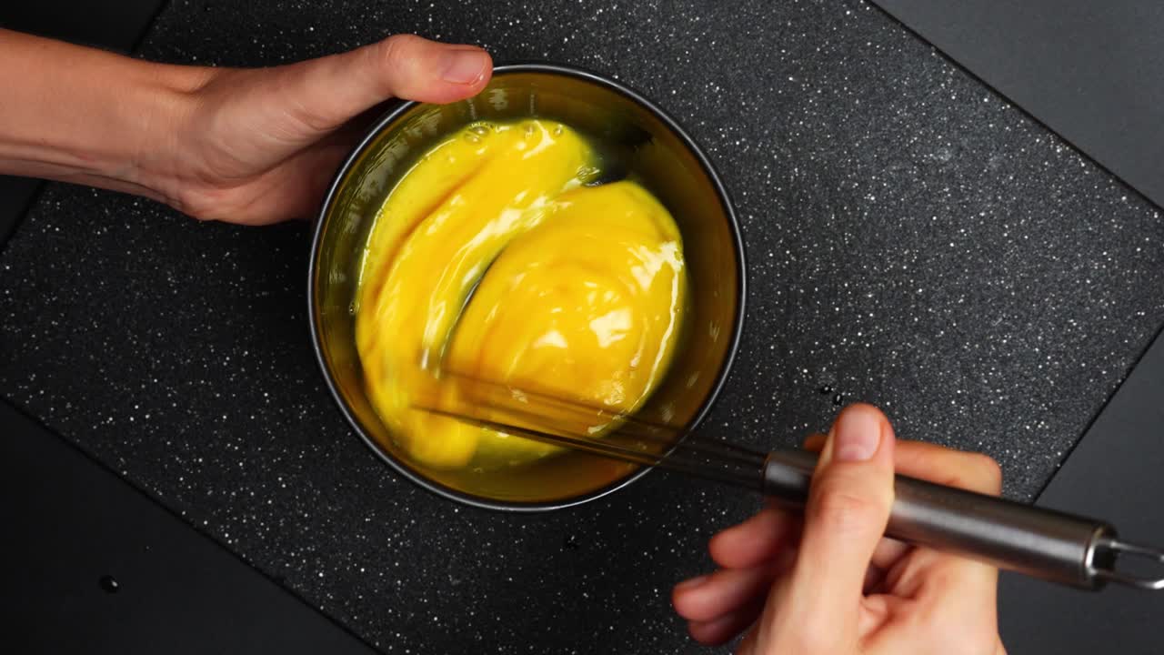 Mengocok telur di mangkuk di atas permukaan gelap. Tangan mengocok telur mentah di mangkuk logam di atas meja dapur gelap