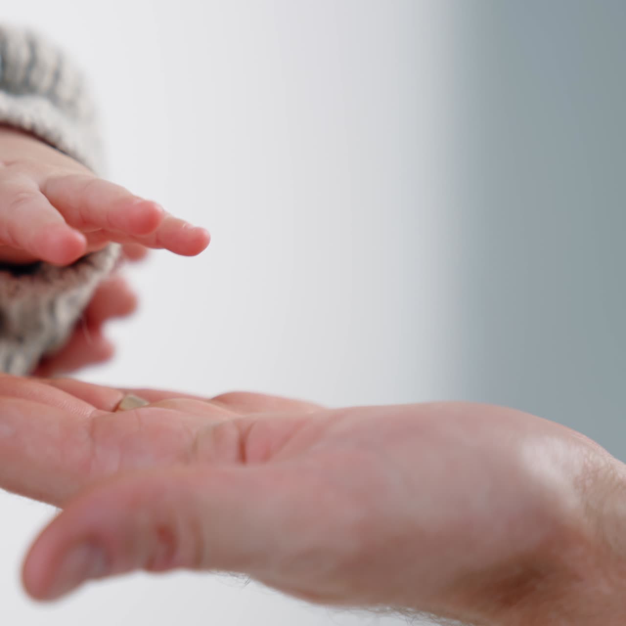 Hand of a little unrecognized baby wearing a grey sweater. Infant's hand is on the male adult hand. Close up. White backdrop