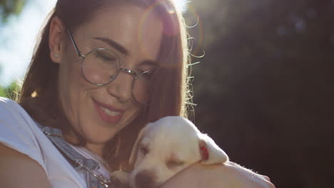 close-up view of caucasian young woman in glasses holding and petting a labrador puppy in the park on a summer day