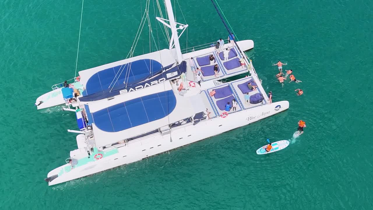 Aerial footage of a catamaran with people swimming nearby in clear, turquoise waters under bright daylight.