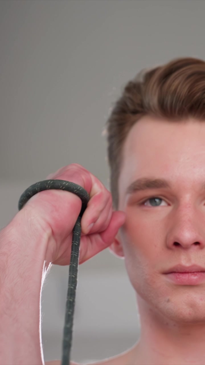 Nærbillede af en ung mand med lysebrunt hår, hvor en anden hånd strammer grebet om et objekt, og hans synlige profil fremhæves. Indendørs omgivelser med neutrale vægge og blød belysning.