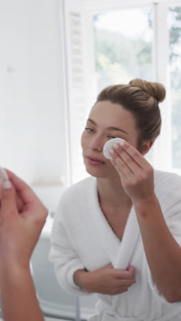 vertical video of biracial woman cleansing face with cotton pad in bathroom, slow motion