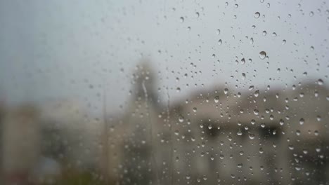 Una-Cámara-Lenta-De-Gotas-De-Lluvia-Vistas-A-Través-De-Una-Ventana-Durante-Fuertes-Lluvias,-Con-Un-Paisaje-Urbano-Al-Fondo