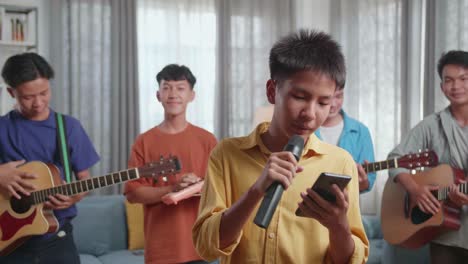 young asian boy in front of his friends with musical instruments holding a microphone and looking at script on smartphone before speaking at home