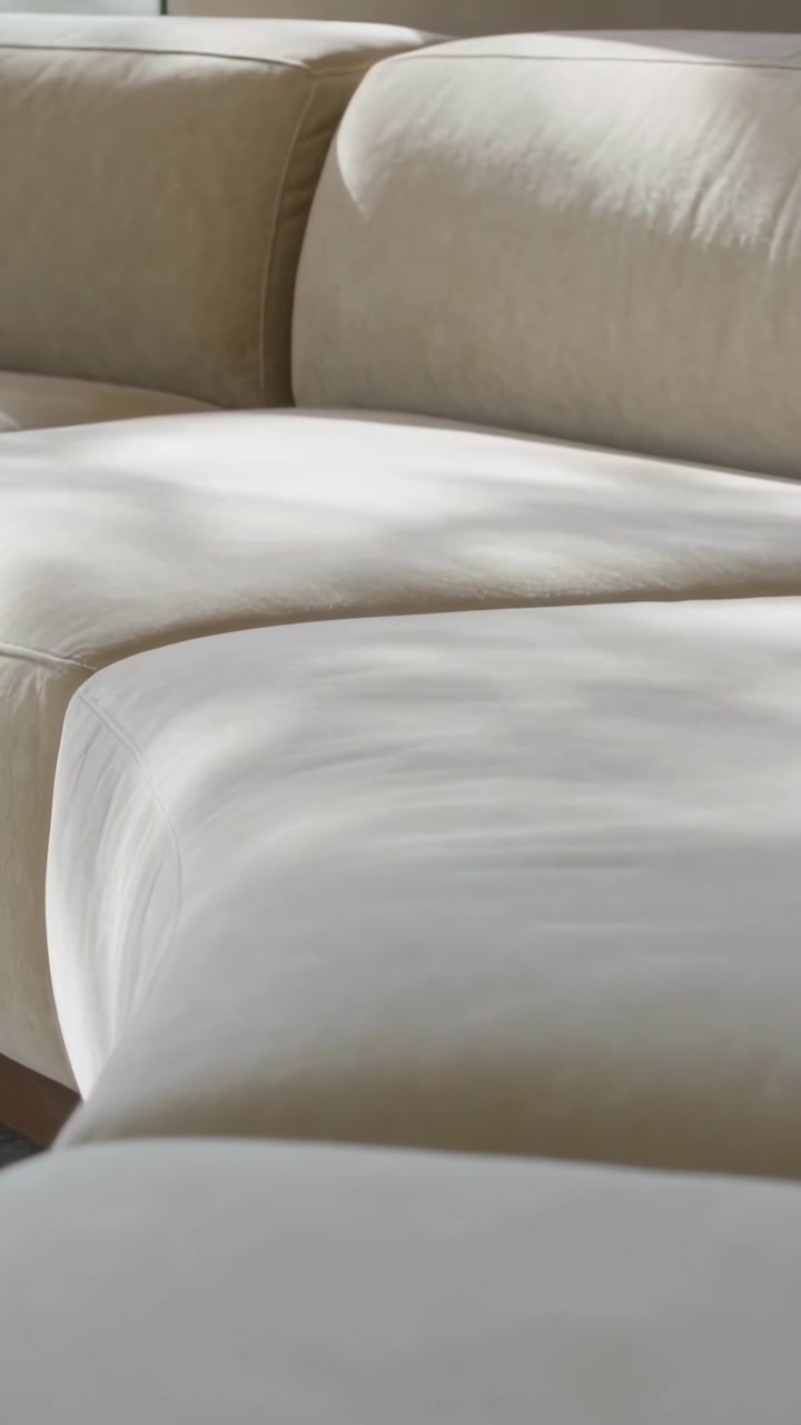 Video verticale: Luce naturale che si sposta dalla finestra proiettando un bagliore soffuso su un divano componibile color crema e un pouf.