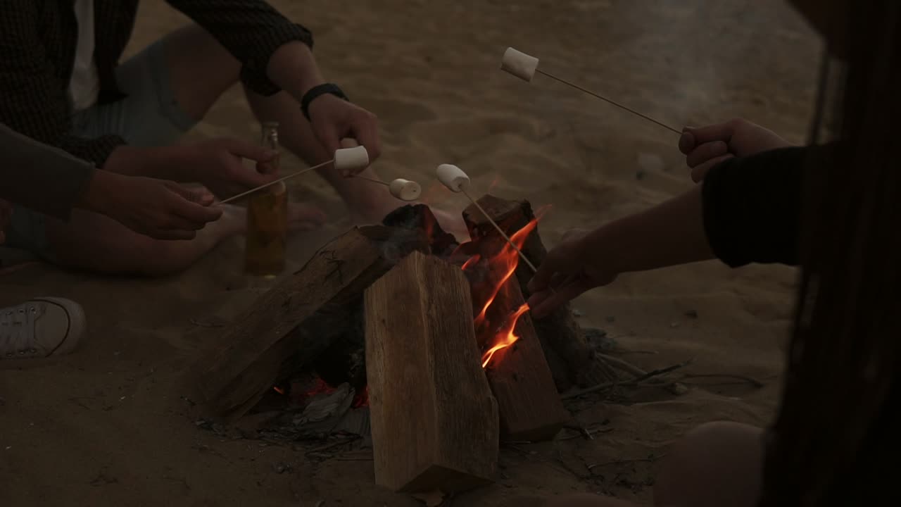 해변에서 불  ⁇ 에 앉아 마시멜로를  ⁇ 고 있는 젊은 친구들의 그룹. 느린 움직임으로 촬영