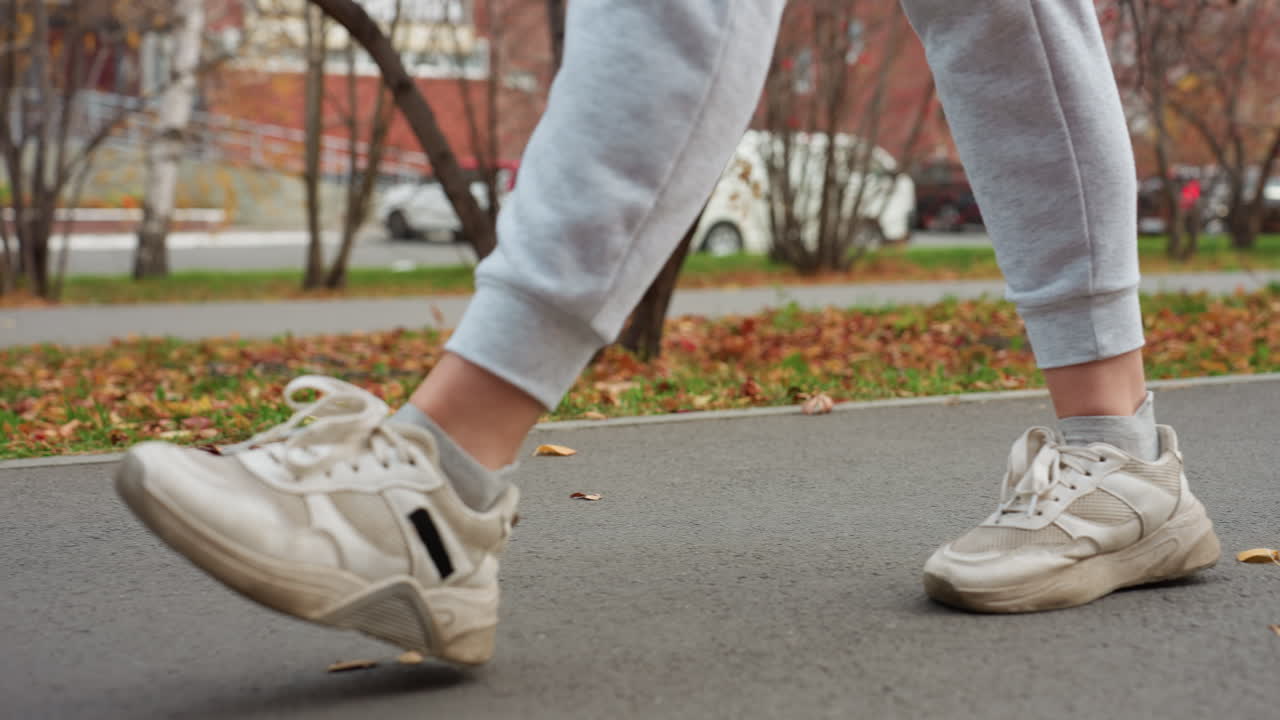 Leg view of person on morning stroll wearing joggers and sneakers walking along marked pathway as white cars remain parked and bus moves in background with autumn leaves scattered on roadside