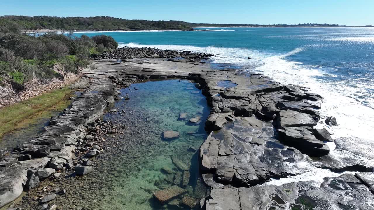 Flyr lavt over en steinstrand, kameraet ser ned i en klar steinkulp som glitrer med sollysrefleksjoner. Blått sommervann skvulper forsiktig mot stein på en lys, skyfri dag. Droneopptak.