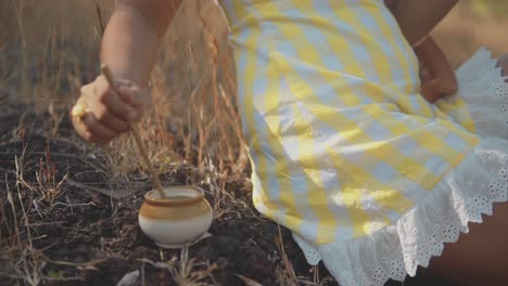 a close up shot of the hand of a female wearing a yellow checkered dress, using a stick slowly stirring a liquid in a clay pot on a dry grass embankment in a grass field, india