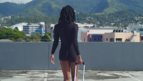 rear view of a young lady walking on a rooftop holding her luggae with mountains in the background