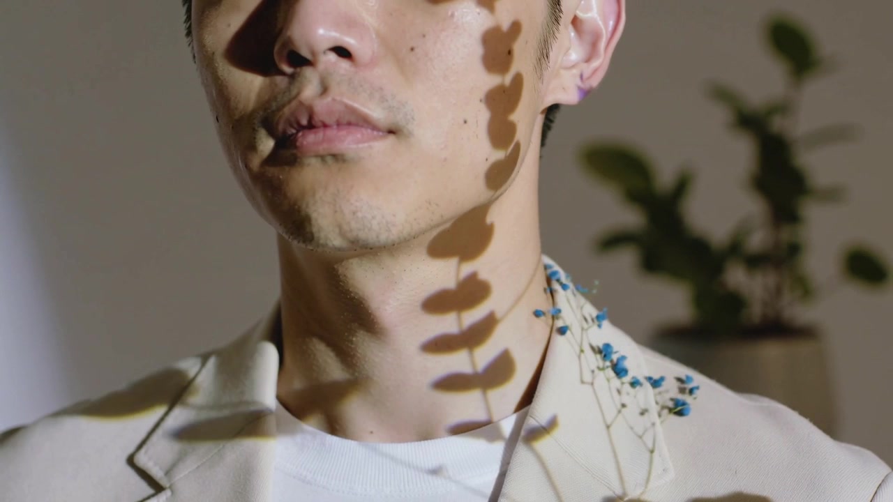 Light starting, casting leaf shadows over male in beige blazer, showing blue blooms in studio