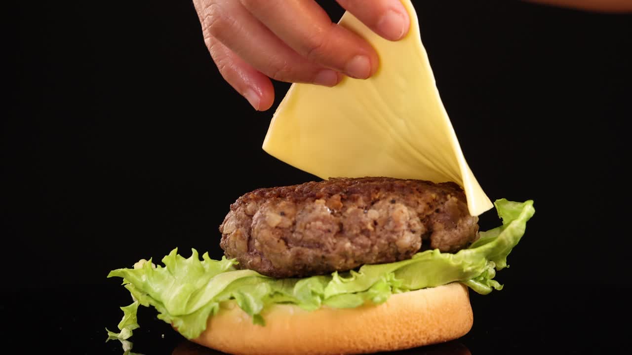 A hand carefully places a cheese slice onto a beef burger patty with lettuce and bun, shot in close-up against a black background with studio lighting