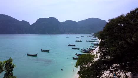 4K-AERIAL-DOLLY-BACKWARD-VIEW-of-a-beach-with-Longtail-boats-moored-in-Phi-Phi-Island-bay,-Phi-Phi-Don,-surrounding-limestone-rocks-cliff-and-the-water-bay