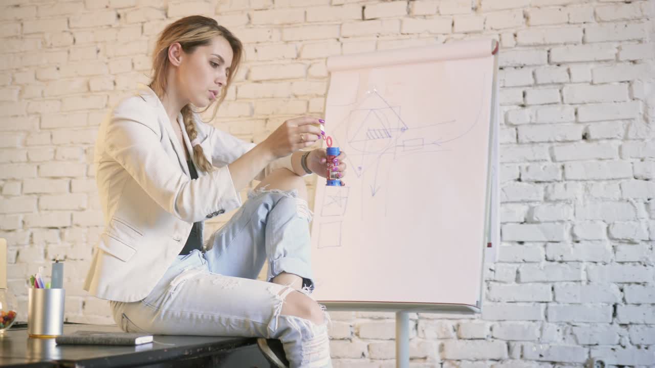 elegante mujer de negocios sentada en el escritorio en la oficina creativa soplando burbujas. vista lateral