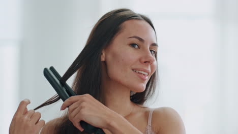 chica feliz usando hierro para el cabello en el baño blanco de primer plano. mujer sonriente peinado
