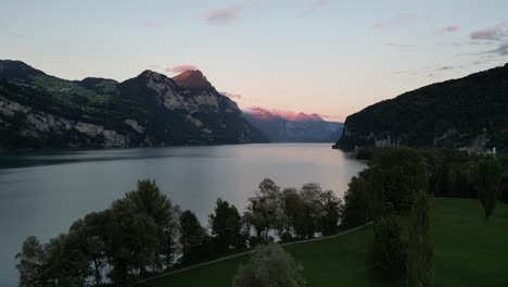 Drone-flight-over-green-meadow-and-trees-against-the-lake-and-mountain-with-beautiful-sky-near-shore-of-Walensee-lake,-Switzerland-in-the-early-morning