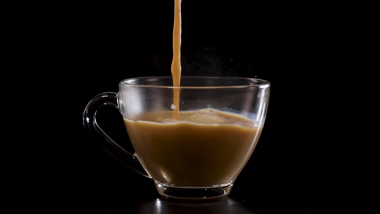 Milk streams into a glass cup of coffee, creating a swirling effect against a dark backdrop