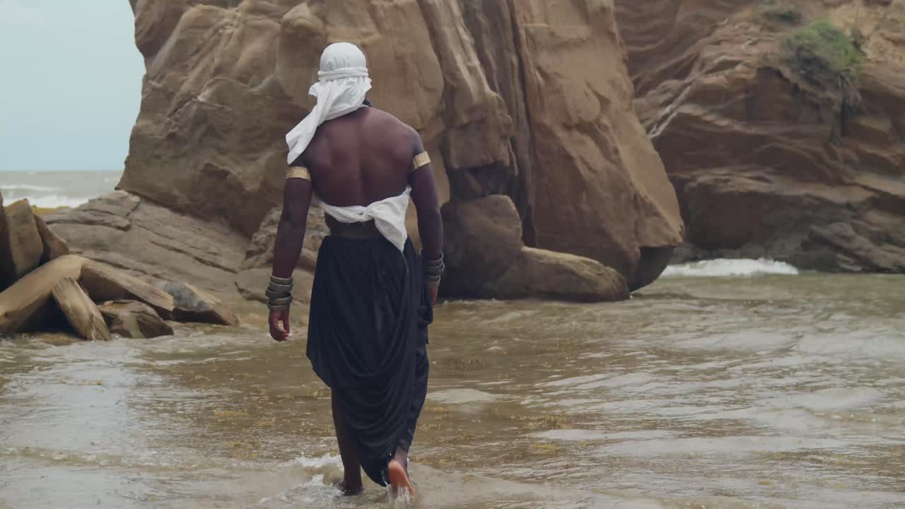 A cosplaying male with dark skin and muscles on the sand with a cliff in the background