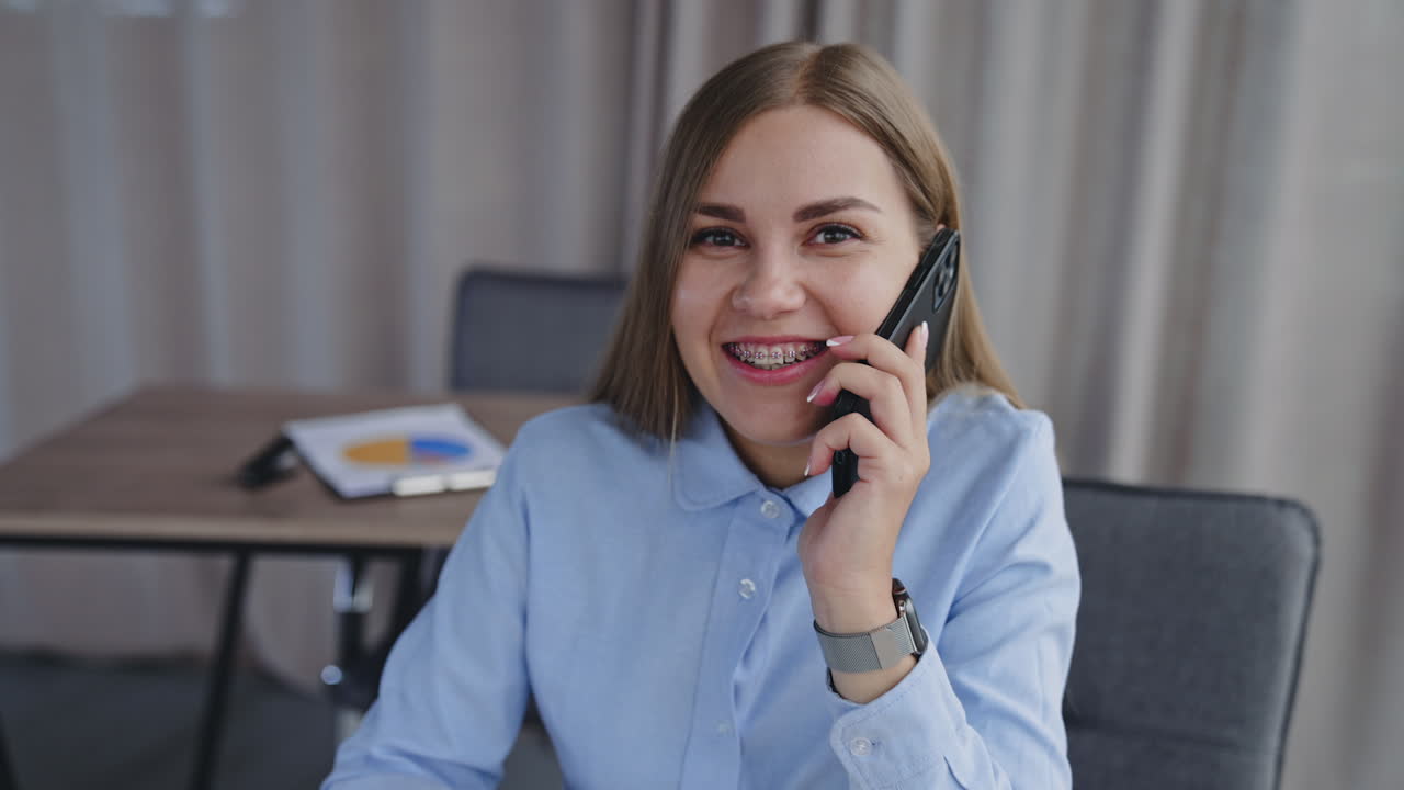 Happy kind smiling woman speaking on the phone sitting in the office. Lady having business phone conversation. Blurred backdrop.