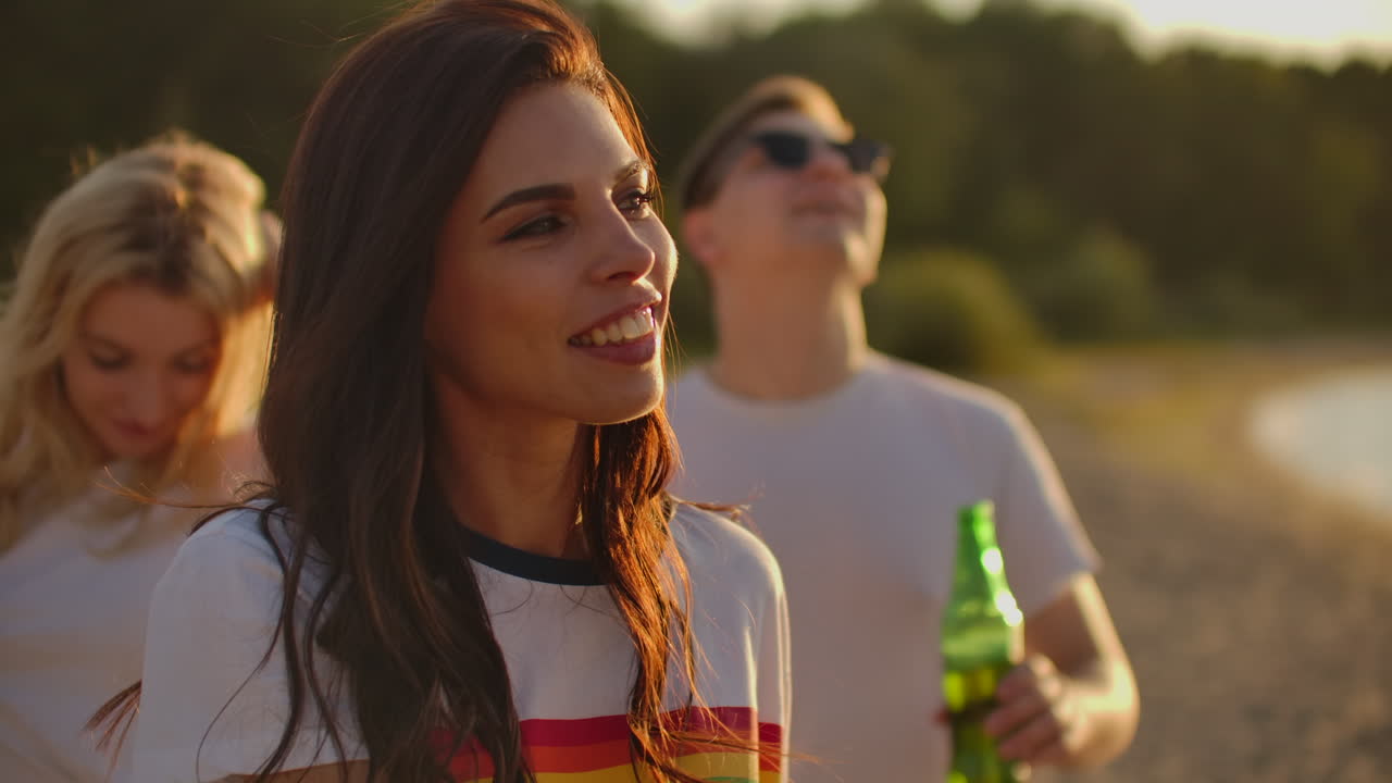 una joven feliz con el cabello largo y oscuro en una camiseta blanca corta está bailando en la fiesta al aire libre con cerveza. ella sonríe y se toca el cabello y disfruta de la fiesta en la costa del lago al amanecer.