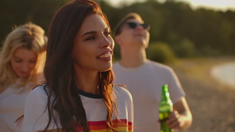 Una-Joven-Feliz-Con-El-Pelo-Largo-Y-Oscuro-Y-Una-Camiseta-Blanca-Corta-Baila-En-La-Fiesta-Al-Aire-Libre-Con-Cerveza.-Ella-Sonríe,-Se-Toca-El-Pelo-Y-Disfruta-De-La-Fiesta-En-La-Costa-Del-Lago-Al-Amanecer.