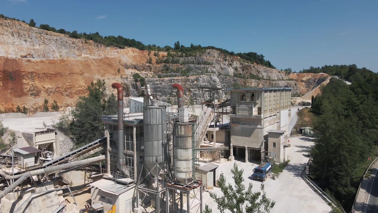Drone video reveals a sprawling quarry and mineral processing plant set against rocky cliffs in Savinjska, Slovenia, on a clear sunny day.