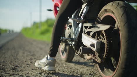 close-up of a biker adjusting the kickstand of a motorbike, with a blurred background showing a car approaching from a distance