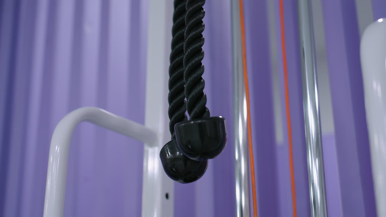 primer plano de equipos de gimnasia colgando de una estación de entrenamiento, el accesorio de cuerda negra contrasta con la elegante estructura de gimnasio blanca y metálica, puesta contra un vibrante fondo púrpura