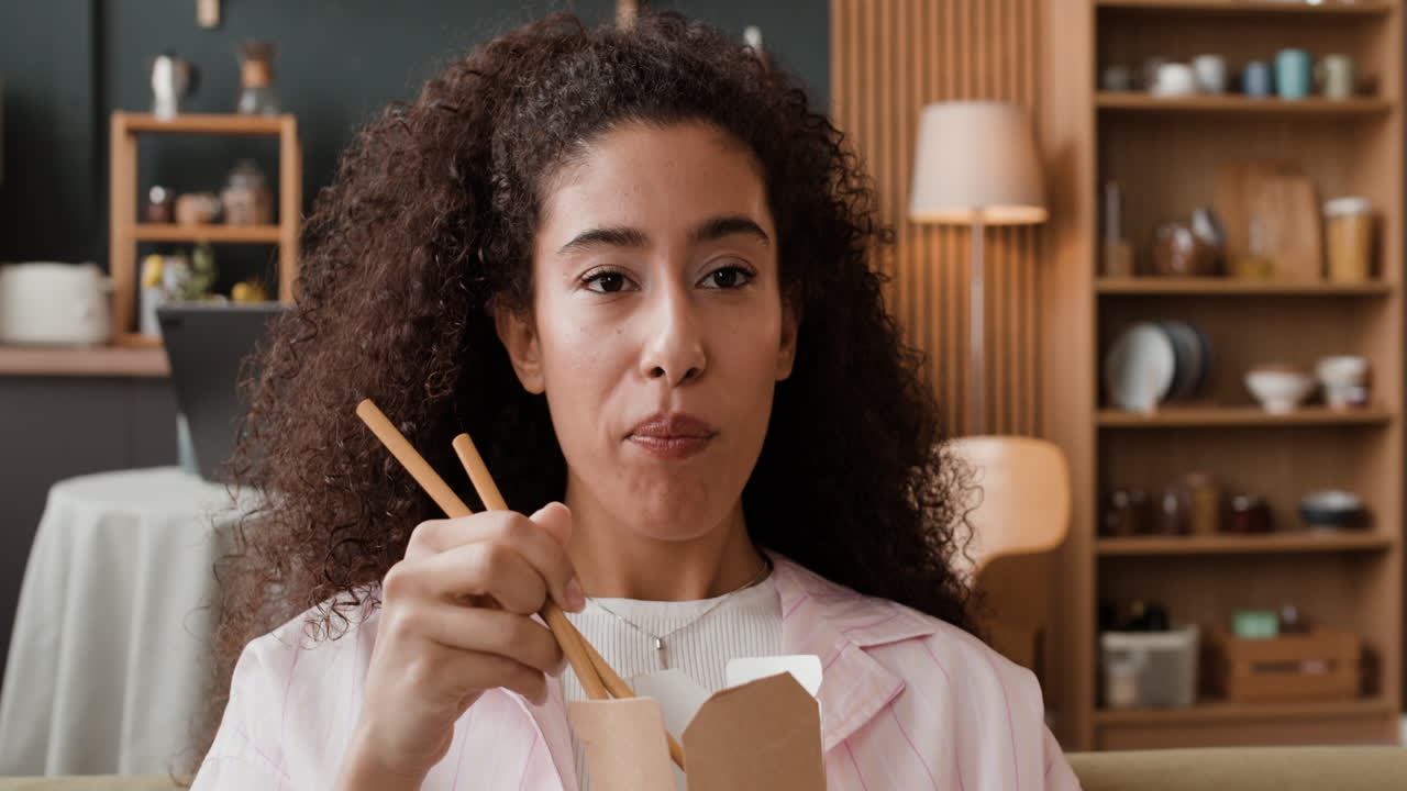 Young woman enjoys eating noodles with chopsticks from a take-out box at home