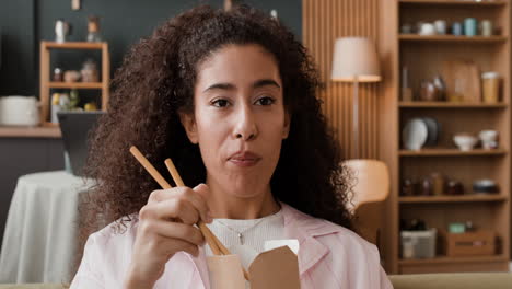 young woman enjoys eating noodles with chopsticks from a take-out box at home