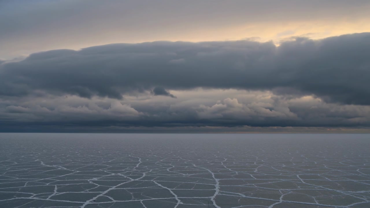 Rippling low cloud bank shifting over cracked frozen ice plain, sun peeking lighting horizon glow