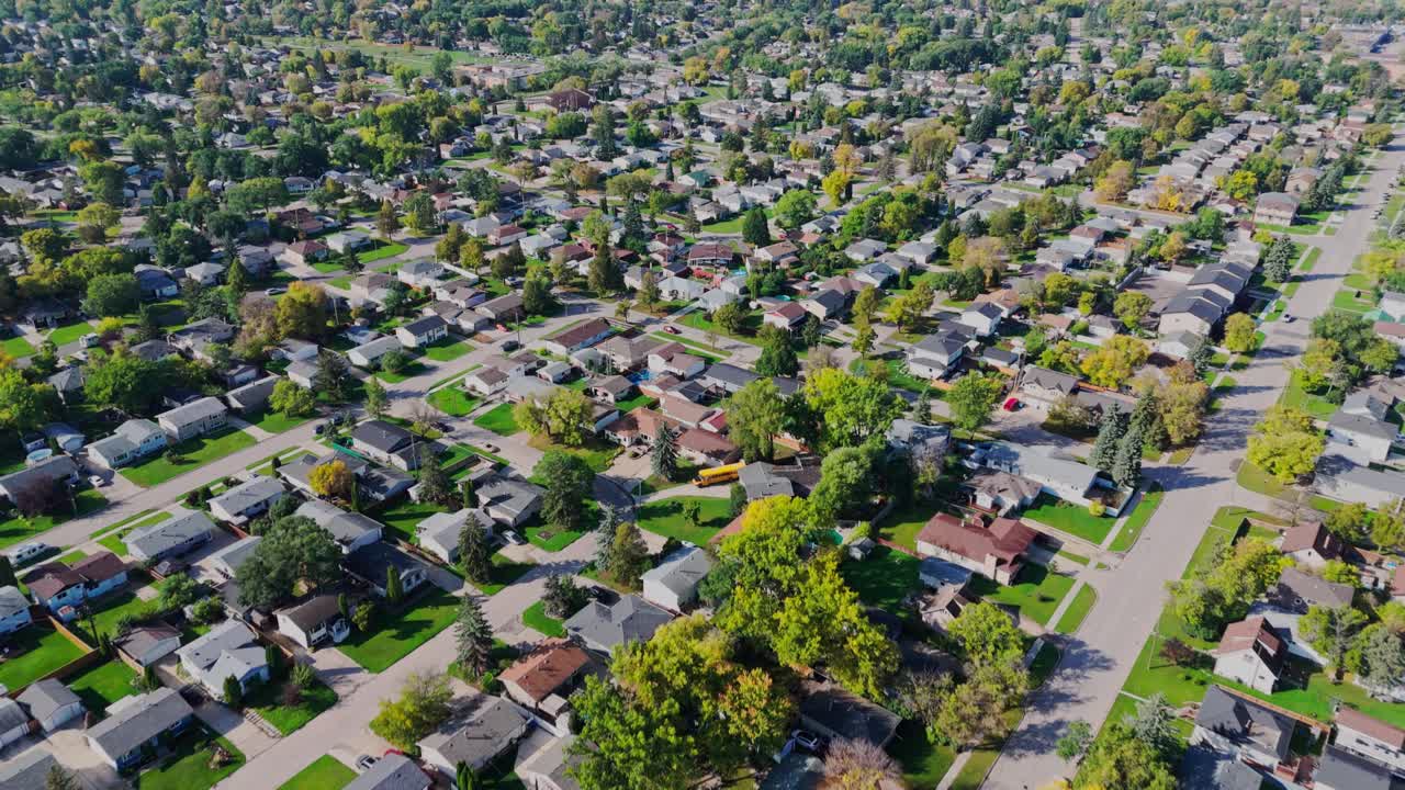 Drone footage of a suburban residential neighborhood featuring rows of houses, streets, and trees with early autumn foliage. A clear view of urban planning and community living