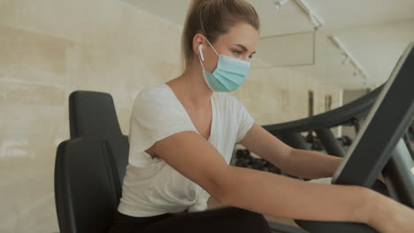 athlete female with face mask working out with an exercise machine and drinking water in the gym