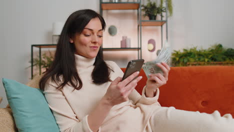 smiling happy young woman at home counting money cash use smartphone income saves lottery win