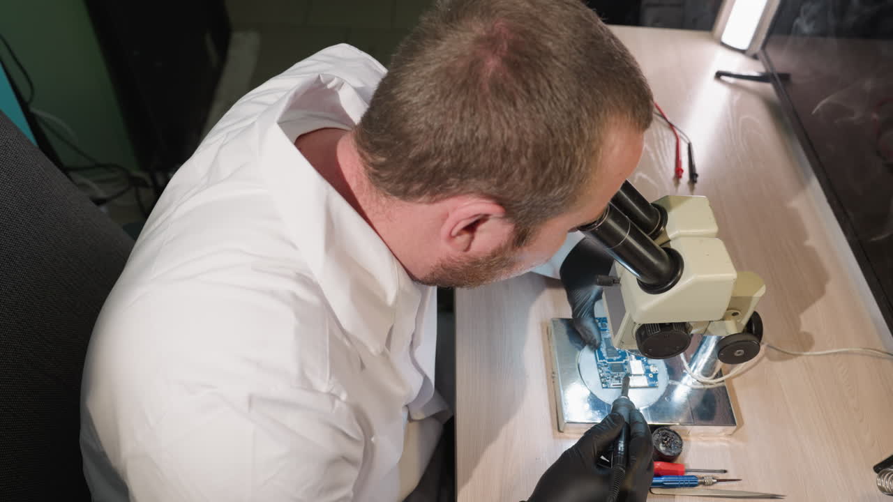 A downward view of a technician in a white lab coat working on a circuit board under a microscope, the workspace is equipped with various tools