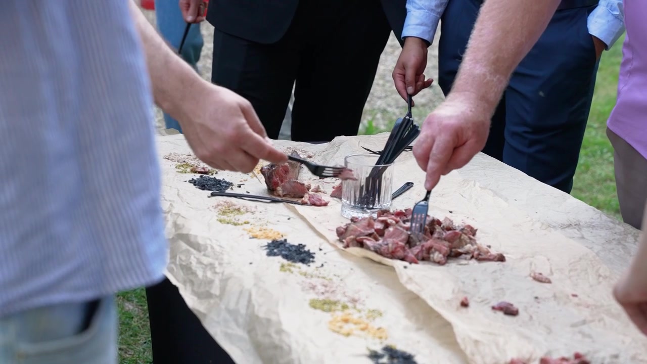 Un gruppo di uomini in abbigliamento casual elegante si riunisce attorno a un tavolo rustico all'aperto, gustando carne tenera e spezie assortite alla luce naturale del giorno, catturando momenti di convivialità vivaci e spontanei
