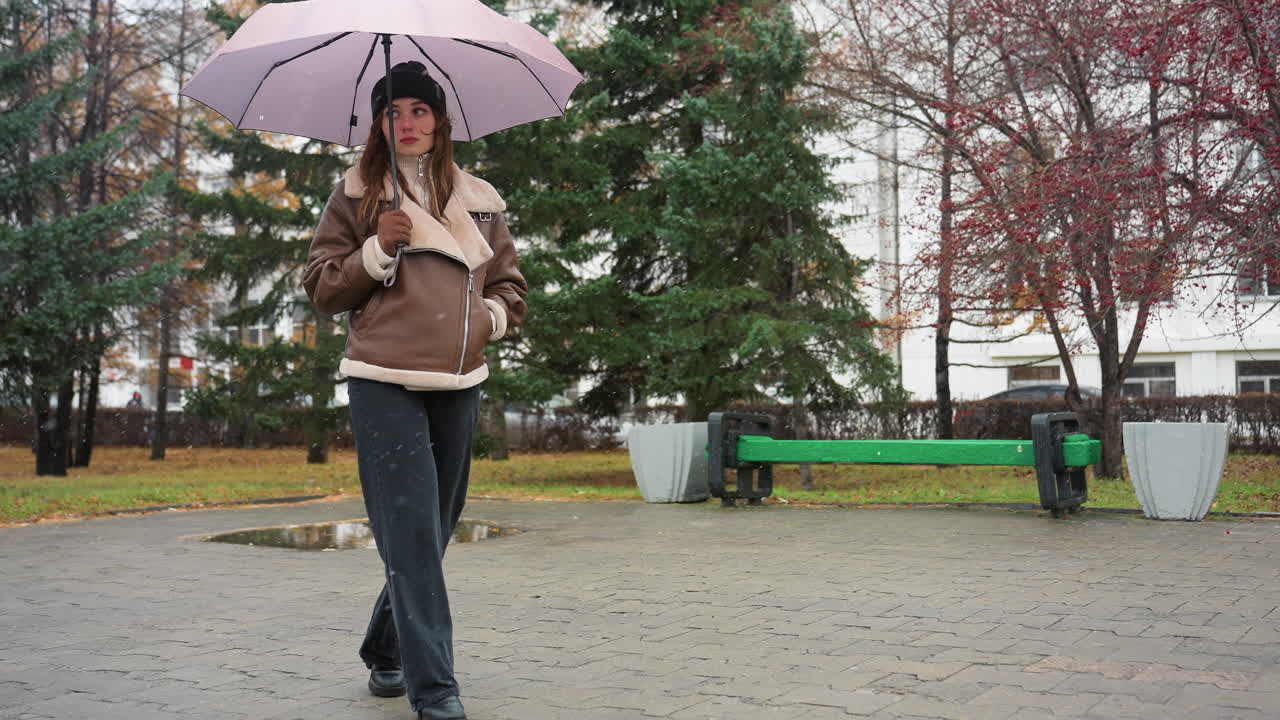 Lady holding umbrella while walking calmly in black knit cap, brown shearling jacket, and black trousers on cold overcast day with light snowfall, surrounded by evergreen trees and wet pavement