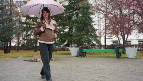 lady holding umbrella while walking calmly in black knit cap, brown shearling jacket, and black trousers on cold overcast day with light snowfall, surrounded by evergreen trees and wet pavement