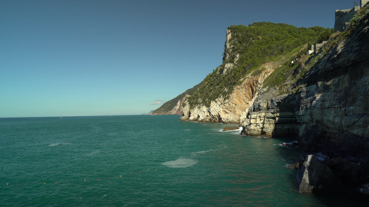 Rocky Ligurian coastline near Porto Venere facing toward Cinque Terre on a clear sunny day, with turquoise Mediterranean sea, steep cliffs, and lush green vegetation along the rugged shore.