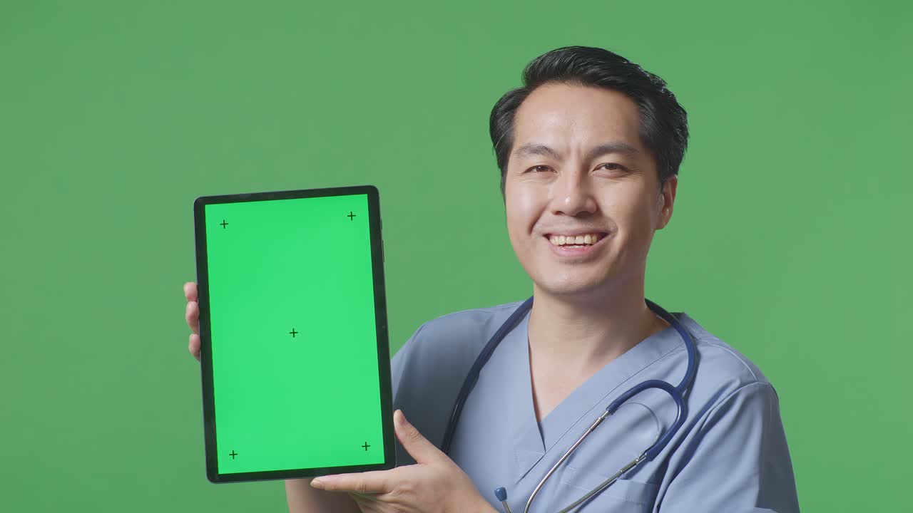 스테토스코프를 들고 있는 아시아 남성 의사의 근접 사진. 병원에서 녹색 스크린 배경에 서 있는 동안 미소 짓고 녹색 화면 태블릿을 보여줍니다.