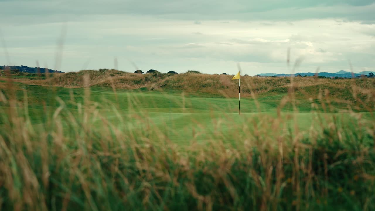 bandera en el campo de golf de ireland links poniendo verde mirando desde detrás de la hierba alta en primer plano áspero, soplando en el viento en cámara lenta